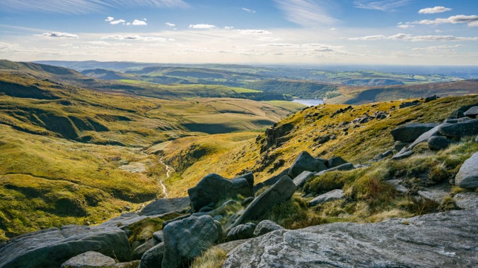 A view from the Pennine Way into a green valley on the High Peak Estate, with rocky terrain in the foreground and running water visible in the valley.